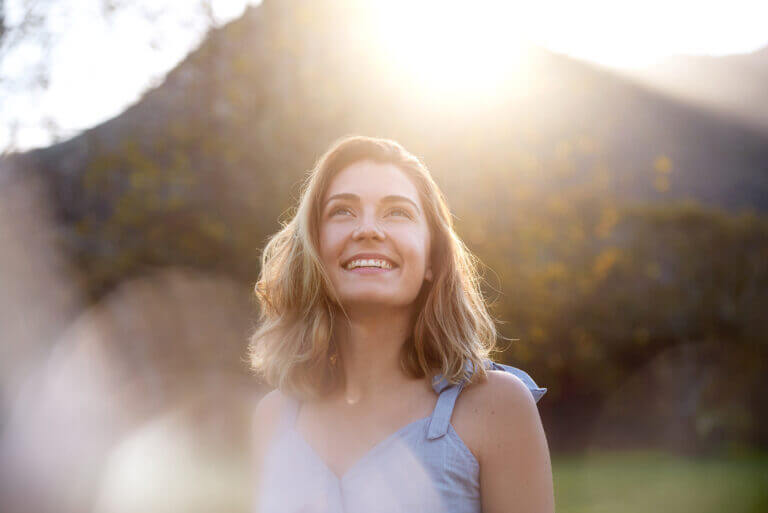 Person standing outdoors in a natural setting with mountains and sunlight in the background.